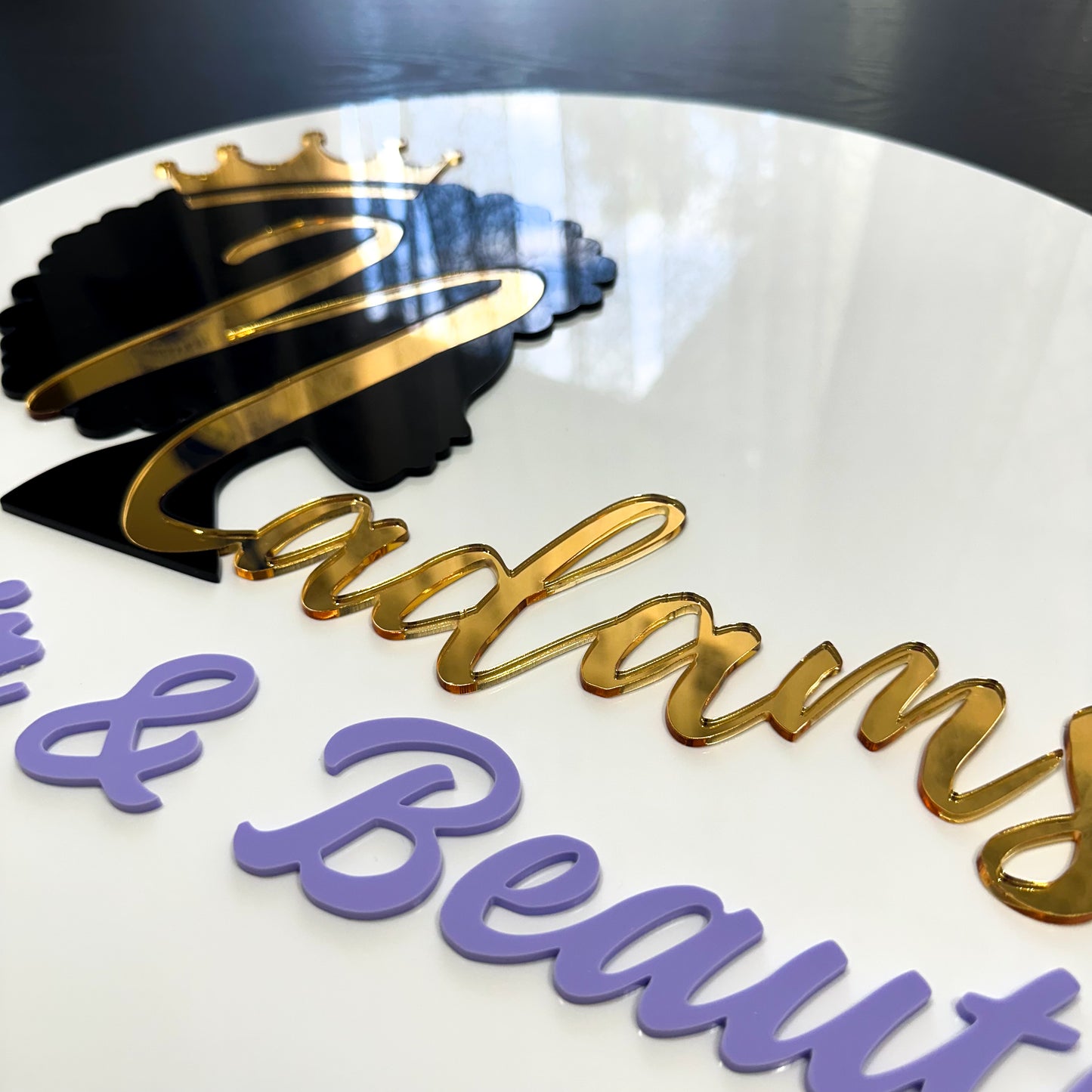 A round acrylic business sign for 'Hair & Beauty' with layered gold, black, and lavender lettering, topped by an ornate crowned silhouette design. The elegant multicolour text contrasts against the white background.