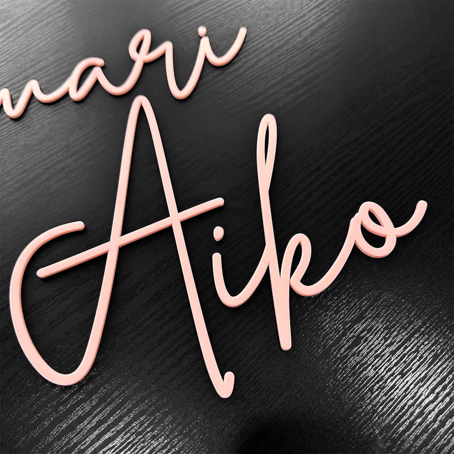A name sign cut from a single layer of baby pink acrylic, with 'Aiko' in lighter pink cursive lettering.